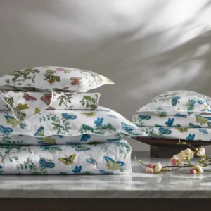 A stack of white pillows and bedding adorned with colorful butterflies and floral patterns, resting on a marble surface. Featuring Baudin Butterfly printed bedding by Matouk, availabe at Linen Society.