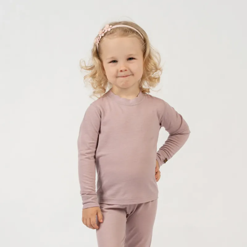 A child with curly blonde hair and a light pink floral headband stands against a white background, wearing a dusty pink long-sleeved top and matching pants. The child has one hand on their hip and is looking at the camera with a slight smile.
