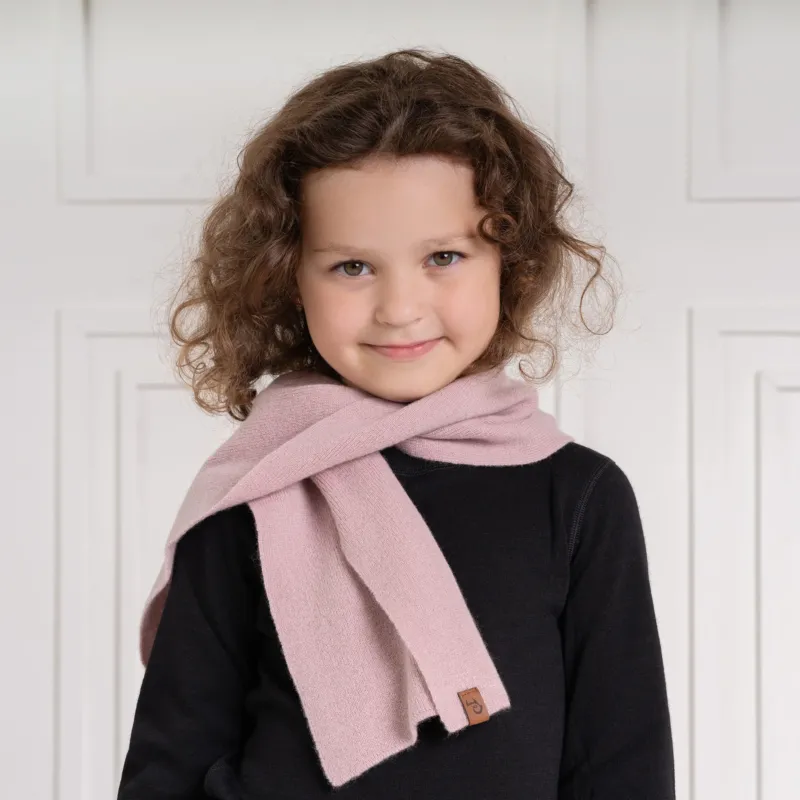 A young child with curly brown hair and a slight smile looks directly at the viewer. They are wearing a black long-sleeved shirt and a light pink scarf with a small brown label, draped around their neck. The background is a white paneled wall.