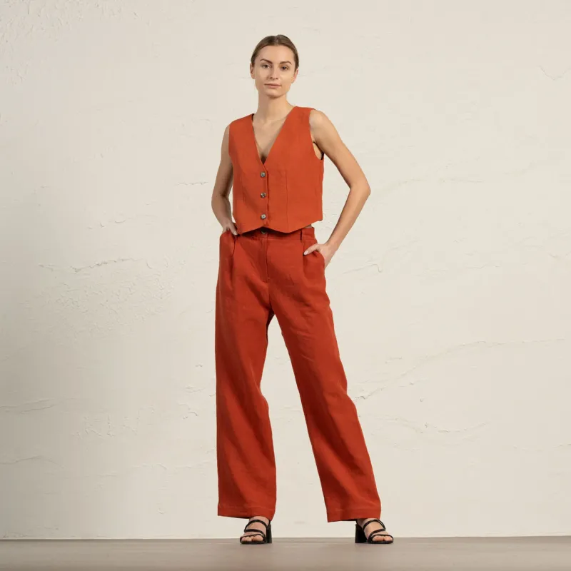 A woman models a cinnamon red linen sleeveless vest and wide-leg pants, standing with hands in pockets against a textured light beige wall. She wears black sandals.