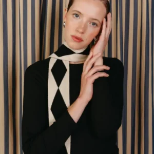 Young woman wearing Merino Wool Skinny Scarf in Black Harlequin pattern, styled with a black top, silver hoop earrings, against a muted striped backdrop.