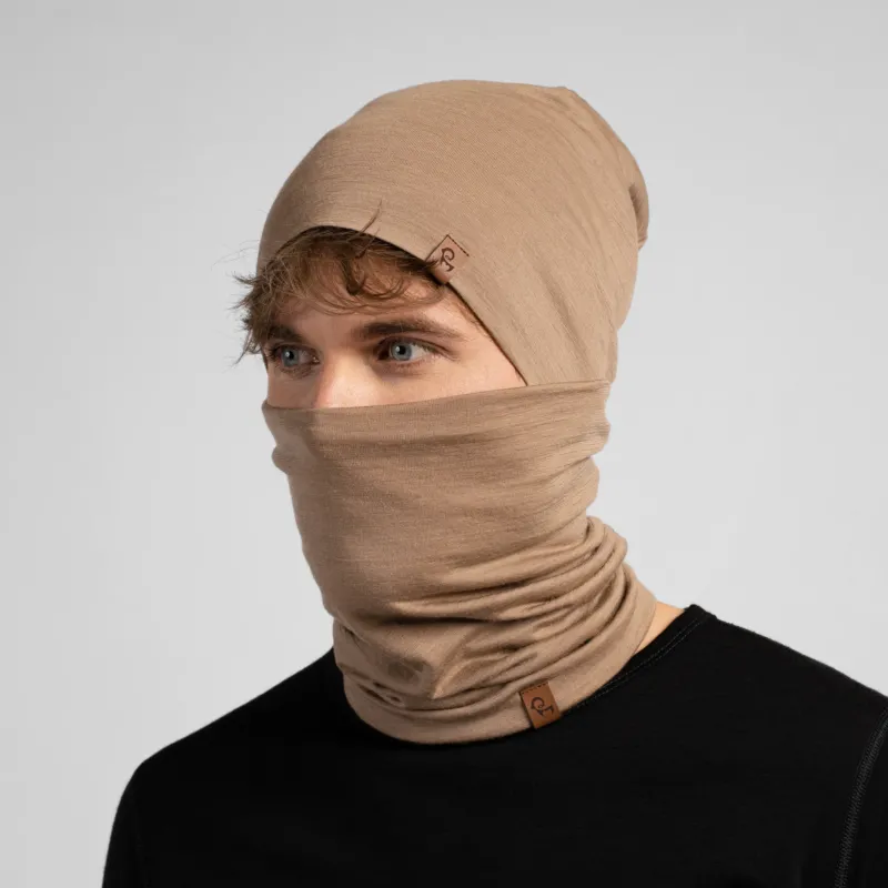 A studio portrait of a man looking thoughtfully off to the left side of the frame. He is wearing both a light beige slouchy beanie and a matching light beige neck gaiter pulled up to cover his nose and lower face. Both items feature a small brown leather logo patch. He is wearing a solid black long-sleeved top against a plain light grey background.