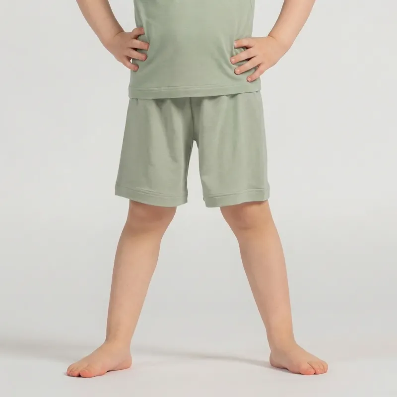 A studio shot, cropped from the chest down, of a young child standing barefoot. The child has their hands resting on their hips. They are wearing a matching set of a light sage green short-sleeved t-shirt and shorts. The background is a plain light grey.