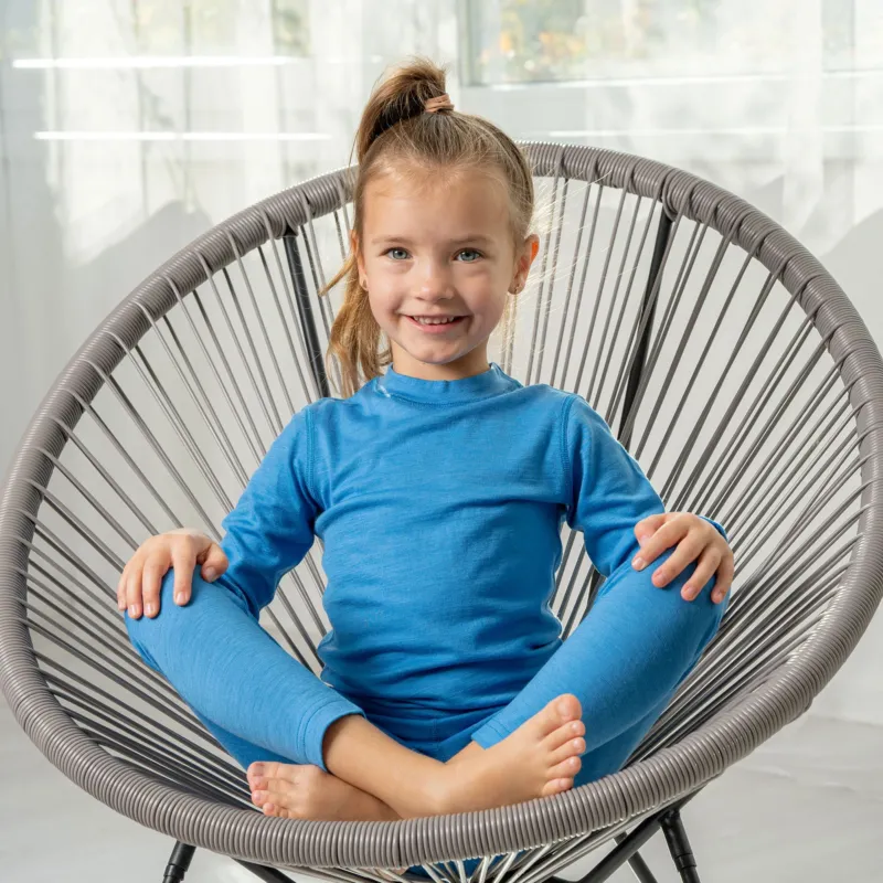 A girl sitting in a longe chair, with her legs crossed and her hands on her knees. She is wearing the merino 160gsm long sleeve top in light blue color and matching pants.