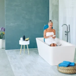 Eine ältere Frau sitzt entspannt in einer freistehenden Badewanne auf einem Badewannenlift mit blauer Rückenlehne und nutzt eine Fernbedienung. Das stilvolle Badezimmer ist modern eingerichtet.