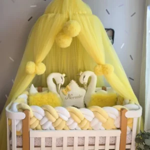 A yellow baby canopy with fluffy pom-poms draped over a white wooden crib, styled with yellow and white braided bedding, two white swan plush toys, and a central pillow that reads 'Nemmin'.