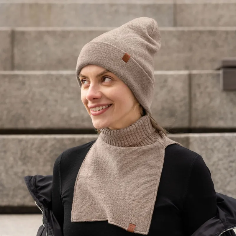 A woman with a fair complexion is pictured outdoors, smiling and looking off-camera to her right. She's wearing a light brown or taupe ribbed knit beanie with a small, square brown logo patch on the left side of the cuff. Around her neck, she has a matching knit neck warmer; this piece features a thick ribbed turtleneck and a rectangular, flat-knit bib that extends down her chest. This neck warmer also has a small brown logo patch at the bottom corner of the bib.
